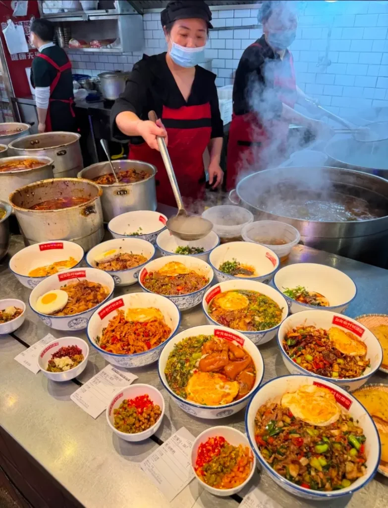 Women preparing Changsha rice noodles at a local breakfast shop, lifting freshly cooked noodles from a pot.